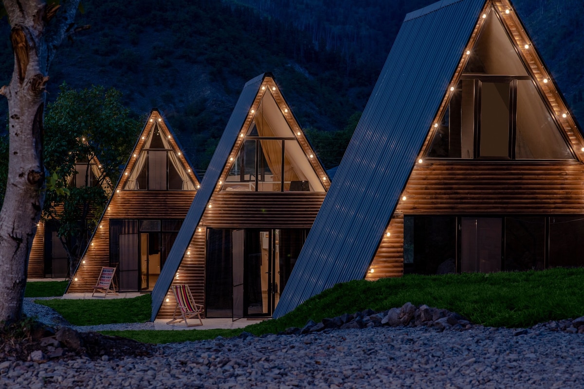A trio of modern cottages with triangular roofs is illuminated by subtle outdoor lighting. Large glass windows reflect the surrounding mountains, while a grassy area is bordered by a stone pathway. Relaxing chairs are visible in front of the cottages, inviting outdoor leisure.