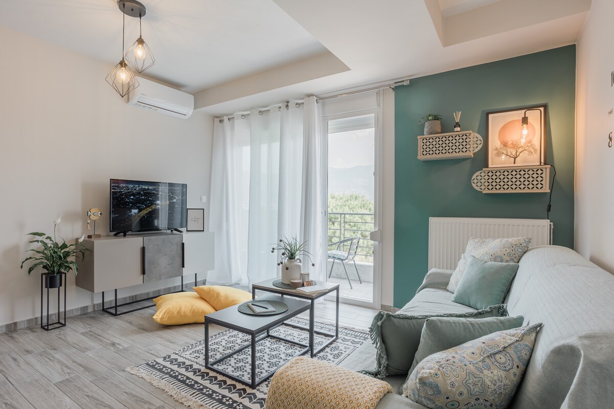 A bright living space features a comfortable sectional sofa adorned with decorative pillows. A sleek flat-screen TV is mounted on the wall opposite, while a modern coffee table sits atop a patterned rug. Natural light fills the room through large balcony doors, enhancing the serene atmosphere.