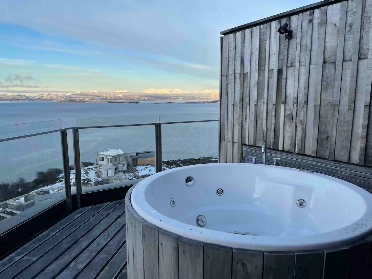 An outdoor jacuzzi is situated on a terrace with wooden decking, offering expansive views of the bay and mountains in the background. The clear water reflects the surrounding landscape, while a glass railing enhances the open feel of the space.