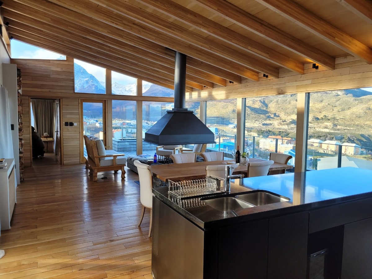 An open-concept living area features large windows that provide panoramic views of the mountains and city. A modern kitchen island is visible, along with a stylish seating arrangement that includes comfortable chairs and a coffee table. Wooden beams enhance the natural appeal of the space.