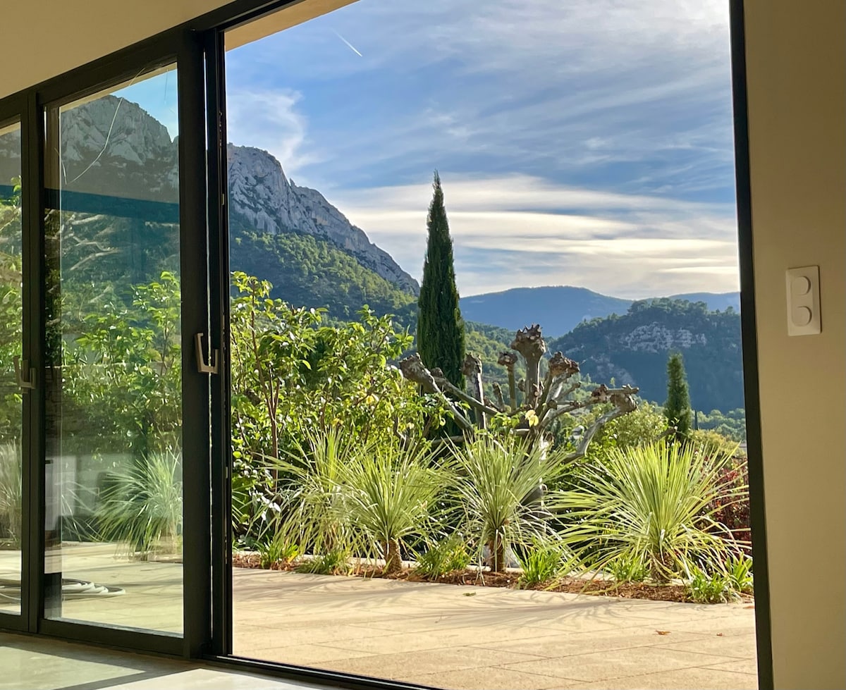 A large glass door opens to reveal a view of lush greenery, with mountains in the background under a clear sky. The outdoor area is surrounded by well-maintained plants, enhancing the natural setting.