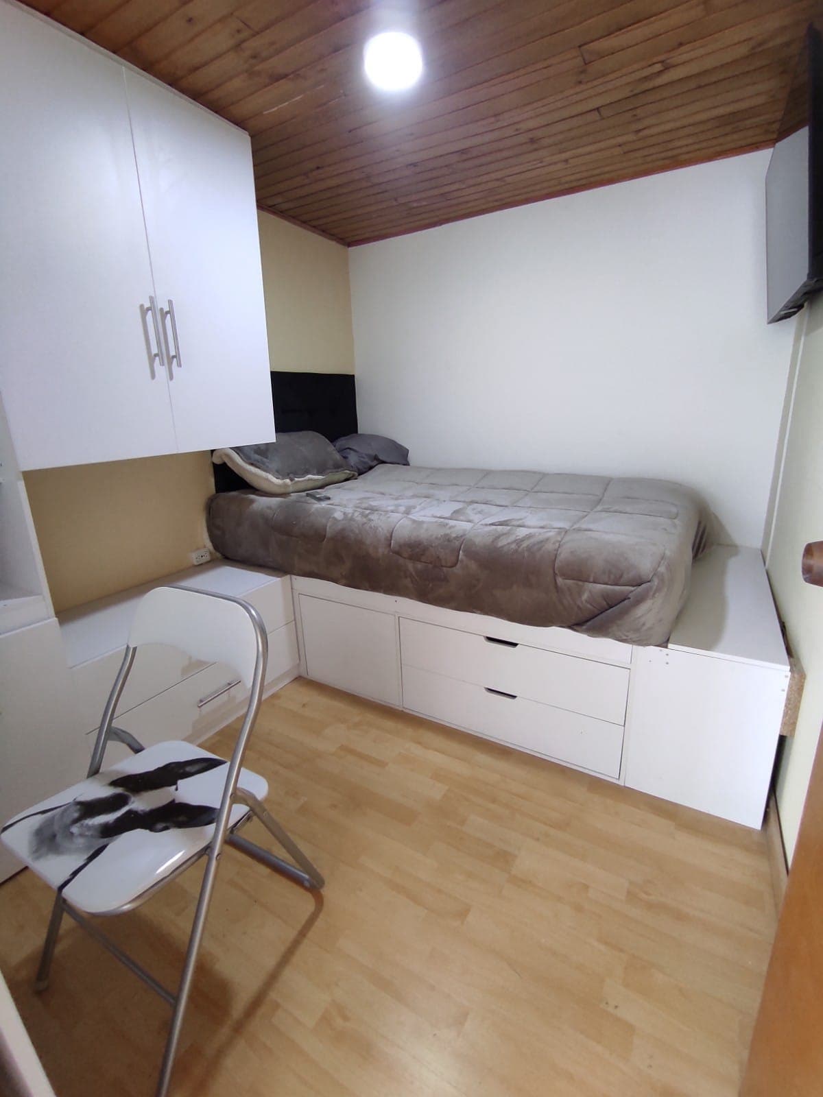 A compact room features a spacious bed covered with a light blanket, positioned against a wall with a television mounted above. A white desk and chair are provided, complemented by built-in storage units below the bed, enhancing the room's functionality.