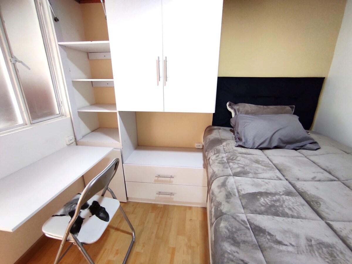 A cozy room features a spacious bed with gray bedding, complemented by a subtle headboard. A desk with a chair is positioned against the wall, while shelves and drawers offer additional storage. Natural light enters through a window, enhancing the room's functionality.