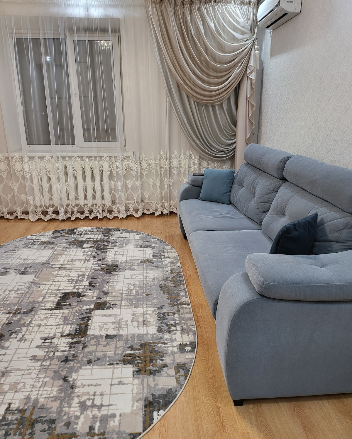 A light-colored, spacious living room features a grey sofa with accent pillows, positioned alongside a patterned oval rug. Large windows draped with sheer curtains allow natural light to fill the space, complemented by a subtle, textured wallpaper.