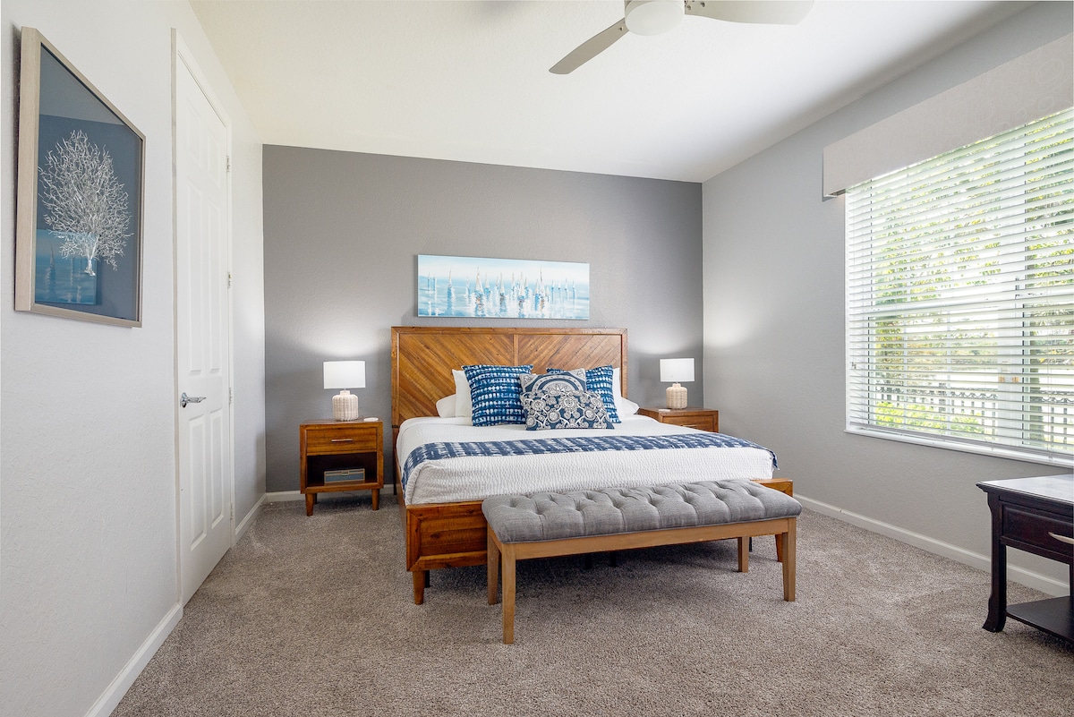 A spacious bedroom features a king-sized bed with new bedding, accompanied by blue decorative pillows. Two bedside tables with lamps stand on either side. A wooden bench is positioned at the foot of the bed. Natural light filters through a window with blinds.