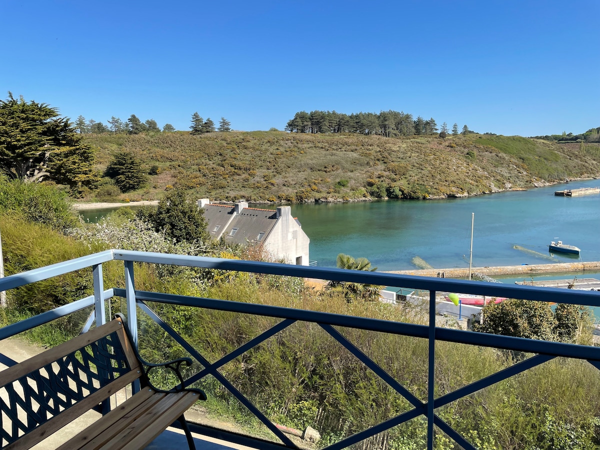 A serene view of the port is presented from a private balcony. Lush green hills rise in the background beside calm waters, while a boat is anchored nearby. A wooden bench frames the scene, inviting relaxation in a peaceful outdoor setting.