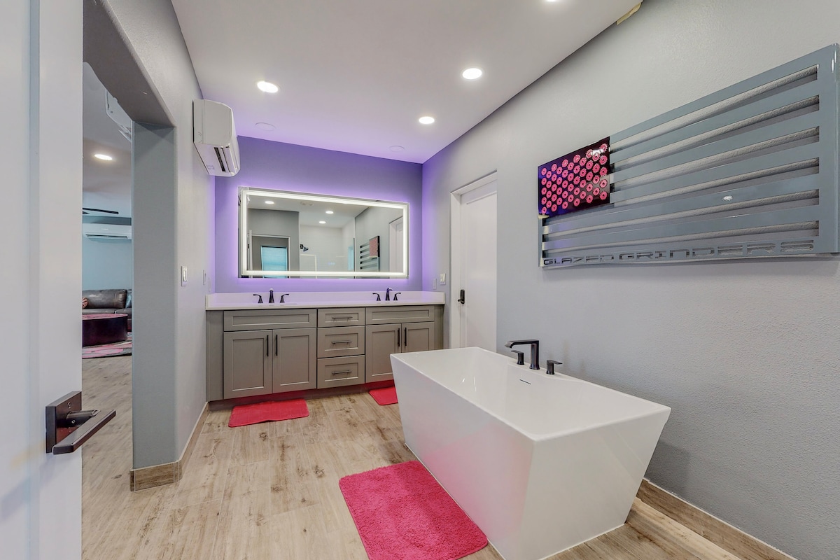 A modern bathroom features dual sinks set within a sleek vanity, with a large mirror illuminated by ambient lighting. A freestanding tub occupies the center, complemented by soft pink rugs on the floor. The space is designed with contemporary finishes and neutral tones.