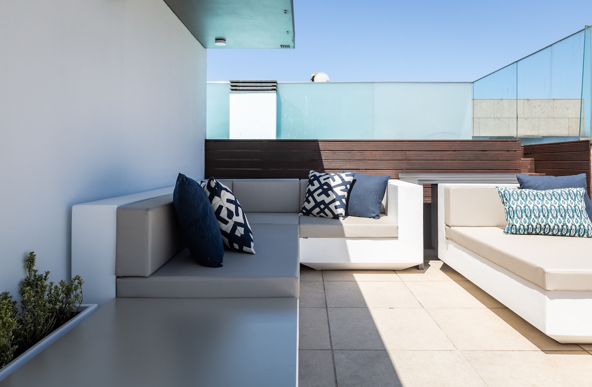 A spacious rooftop terrace is presented, featuring modern seating arrangements. Cushions in various patterns provide comfort on minimalist white furniture. The terrace is enclosed with glass panels, allowing for unobstructed views while providing privacy. Sunlight brightens the tiled floor, enhancing the outdoor sitting area.