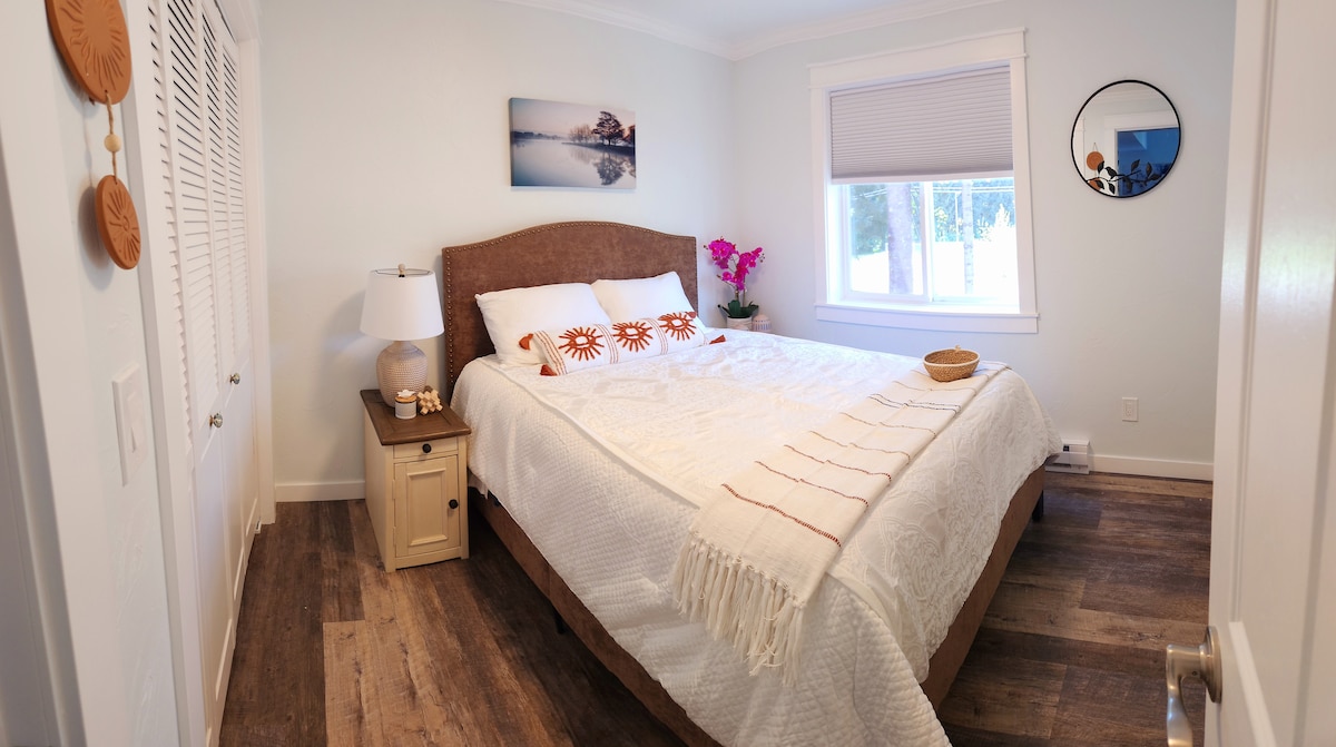 A well-appointed bedroom features a spacious bed with a soft blanket draped across it. A nightstand holding a lamp complements the decor. Natural light enters through a large window adorned with a blind, while a piece of wall art and a circular mirror enhance the room's ambiance.