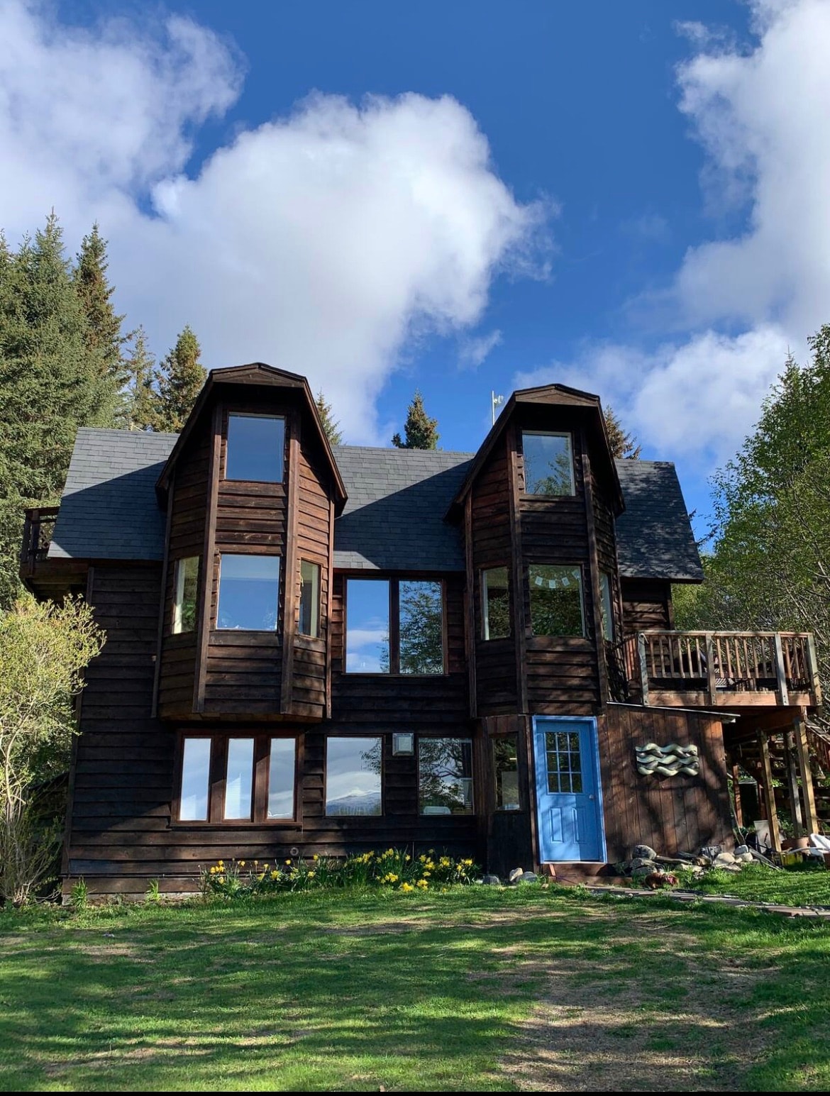 A two-story dark wooden house is surrounded by greenery, featuring multiple large windows that offer natural light. A blue door contrasts with the exterior, and a small balcony complements the architecture. The outdoor area includes a grassy lawn dotted with yellow flowers.