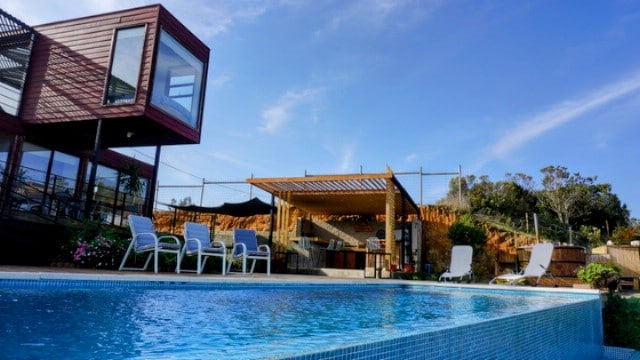 A semi-heated swimming pool is featured in the foreground, surrounded by lounge chairs. In the background, a shaded outdoor seating area is visible, offering a space for relaxation. The landscape includes hills and greenery under a clear blue sky.
