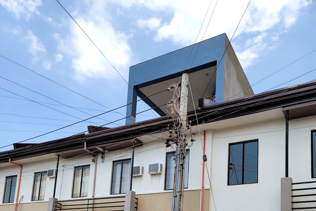 A three-story townhouse is depicted, with modern architecture visible on the upper level. Power lines intersect the sky, while neutral-colored walls and numerous windows are observed on the lower floors, offering a glimpse of the living spaces within.