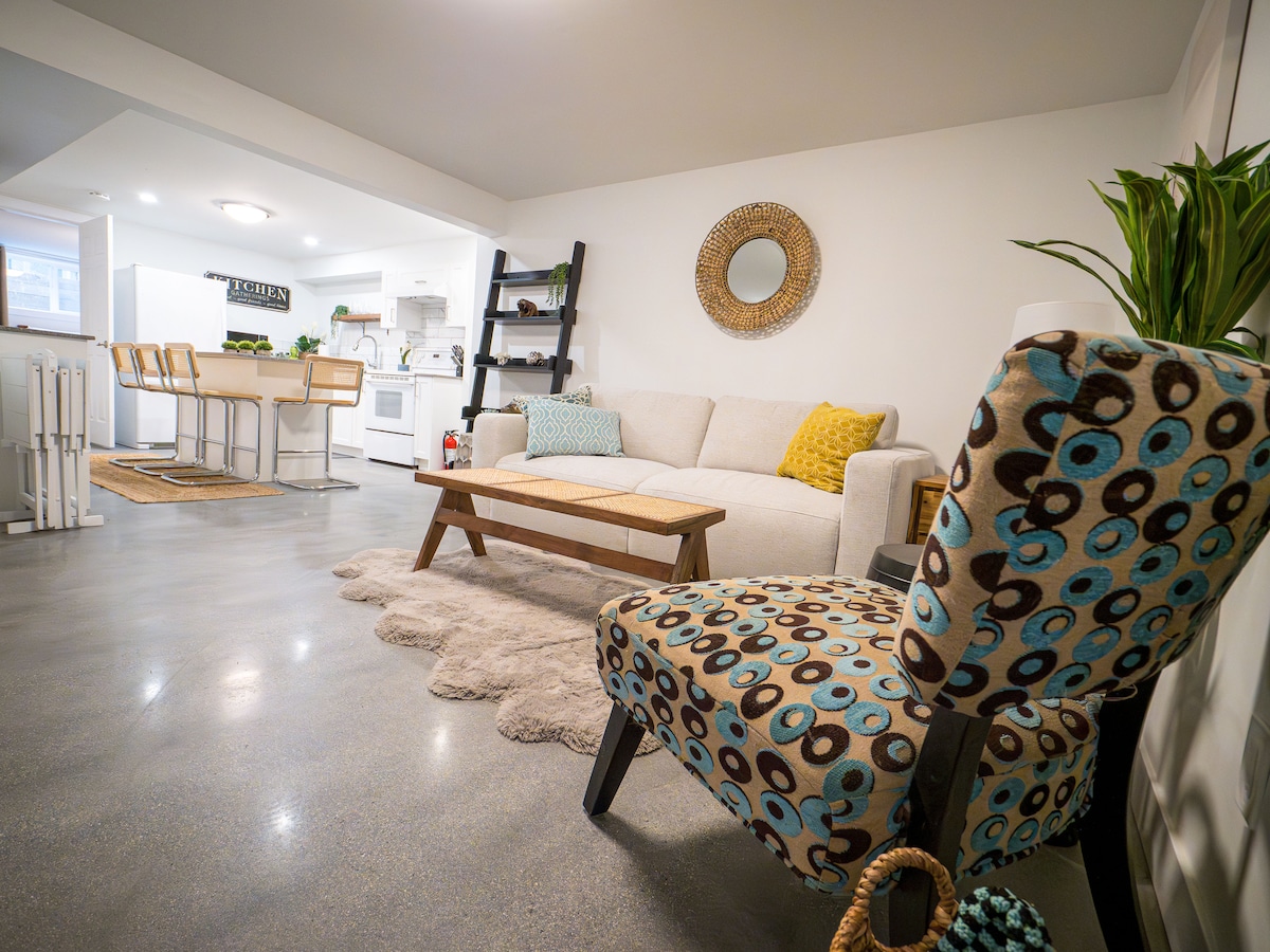 A bright, open living area features a cozy sofa adorned with throw pillows and a textured area rug. A colorful upholstered chair adds a playful touch, while a circular mirror reflects light. A dining space with bar stools and a sleek kitchen is visible in the background.