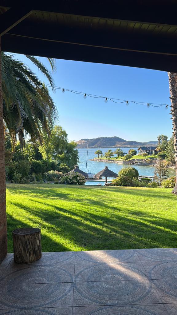 A view from a covered patio reveals a lush green lawn leading down to a serene lake. Palm trees provide shade as the gentle sunlight casts shadows across the yard. A cozy seating area is visible, along with decorative thatched-roof structures on the water's edge.
