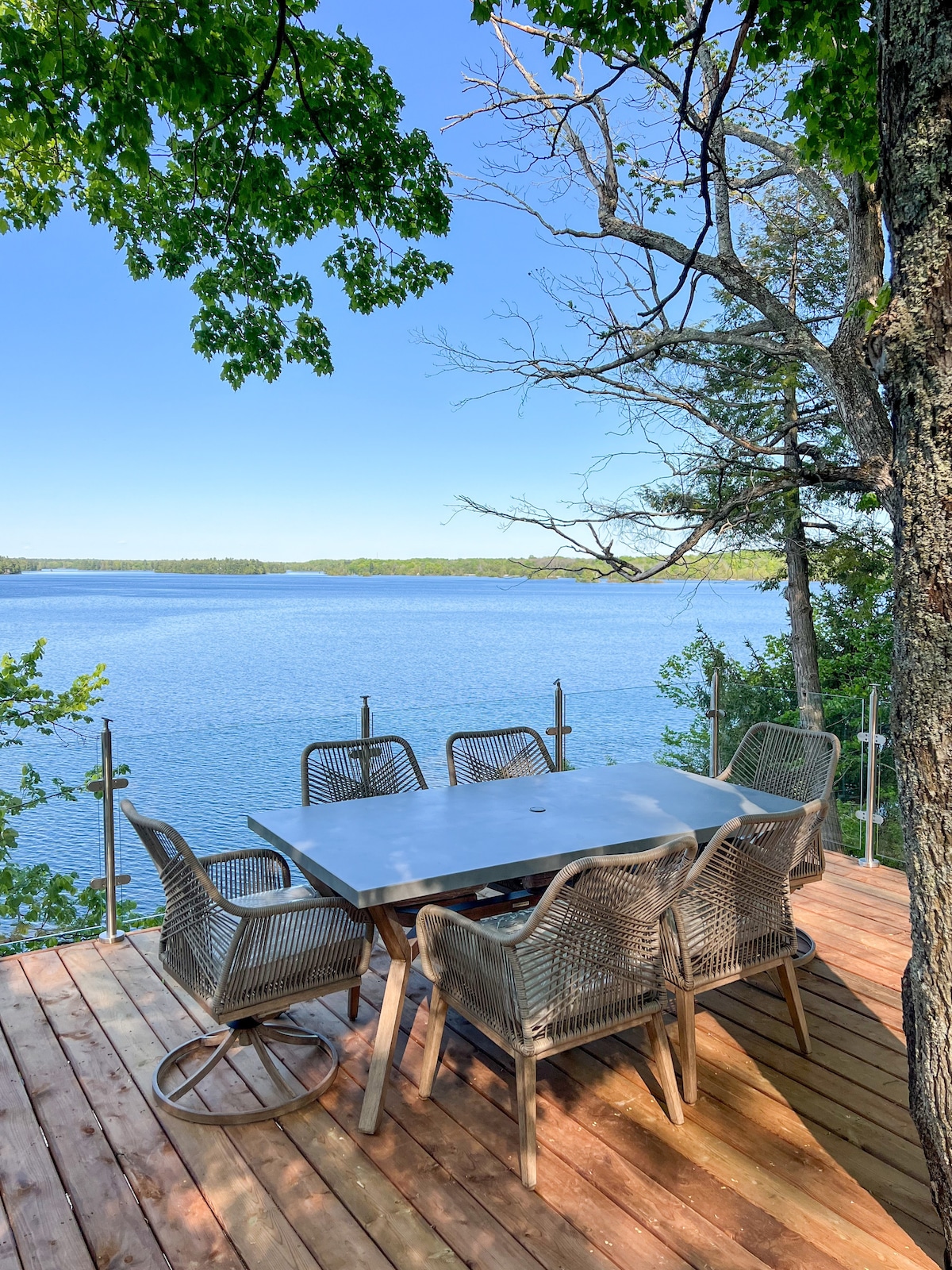 An outdoor dining area is situated on a wooden deck, featuring a large table surrounded by six wicker chairs. The deck offers clear views of the serene lake, framed by lush greenery and tall trees, creating a peaceful setting for meals.