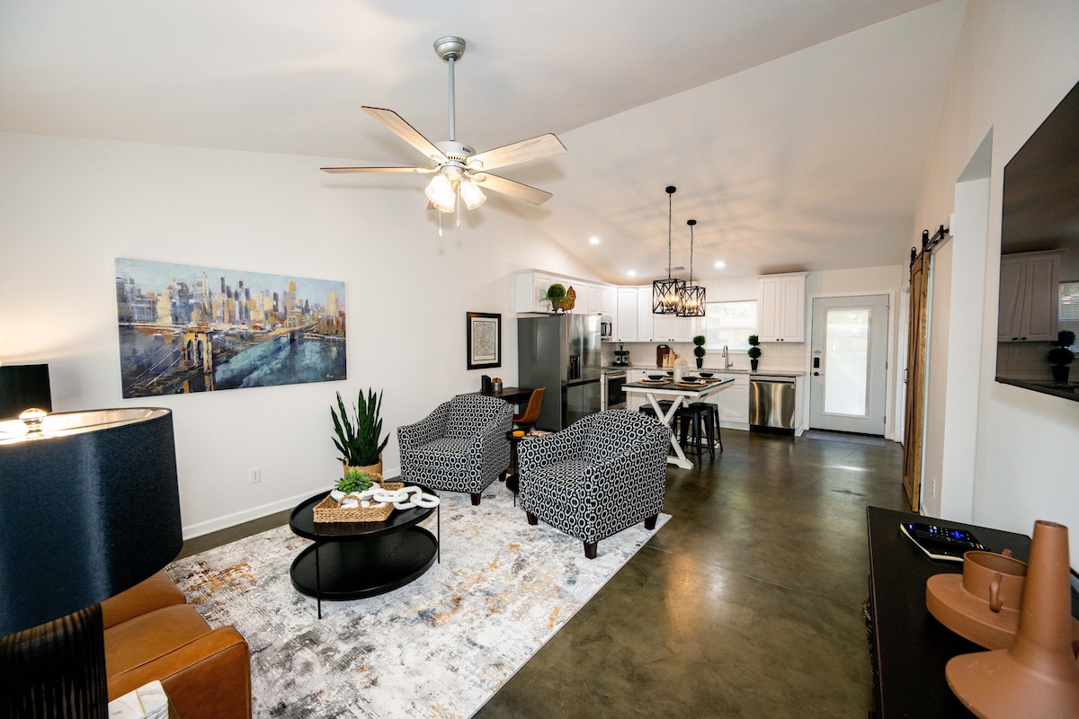 An open living area features two patterned armchairs facing a round coffee table atop a light rug. A modern ceiling fan hangs above, and the kitchen includes stainless steel appliances. Natural light enters through a nearby door, enhancing the space's inviting design.