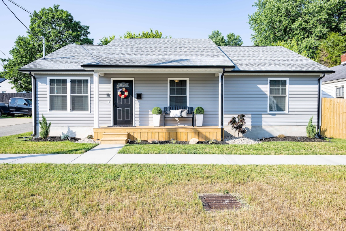 The exterior of a contemporary single-story house is captured, featuring light gray siding and a charming front porch with decorative plants. Green grass surrounds the home, and a well-maintained sidewalk leads to the entrance, which is marked by a welcoming door and a tasteful wreath.