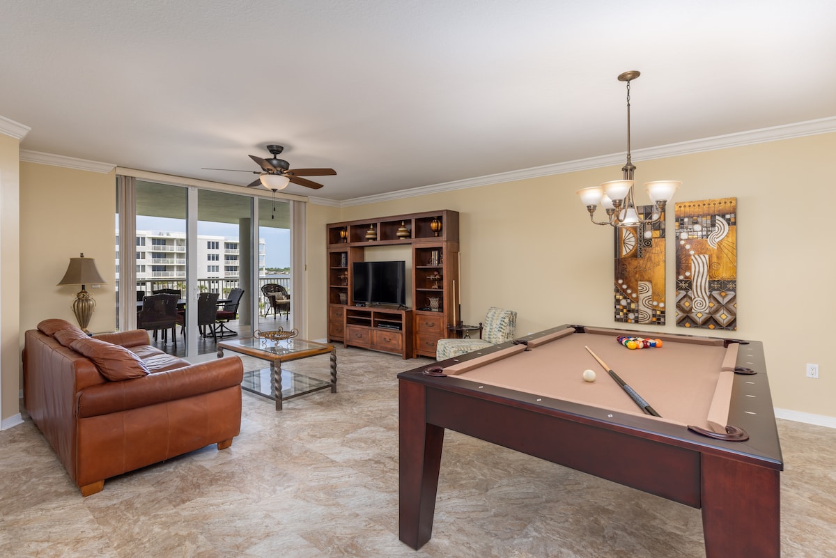 A spacious living area includes a leather sofa and coffee table, with a large TV positioned on a wood cabinet. A 7' slate pool table occupies one corner. Large glass doors lead to a balcony, providing natural light and views of the surroundings.