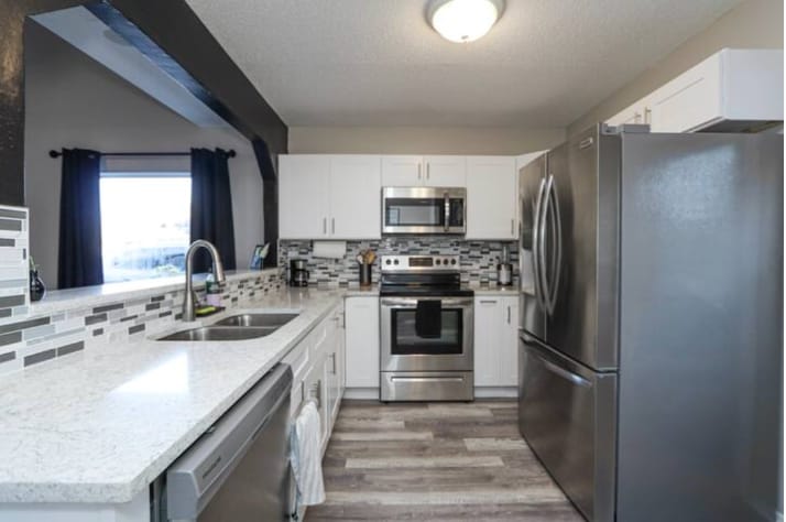 A modern kitchen is equipped with stainless steel appliances, including a refrigerator and oven. A silver sink and a double countertop provide ample workspace. Mosaic tile backsplashes in neutral tones add texture, while natural light is permitted through a nearby window covered by dark curtains.