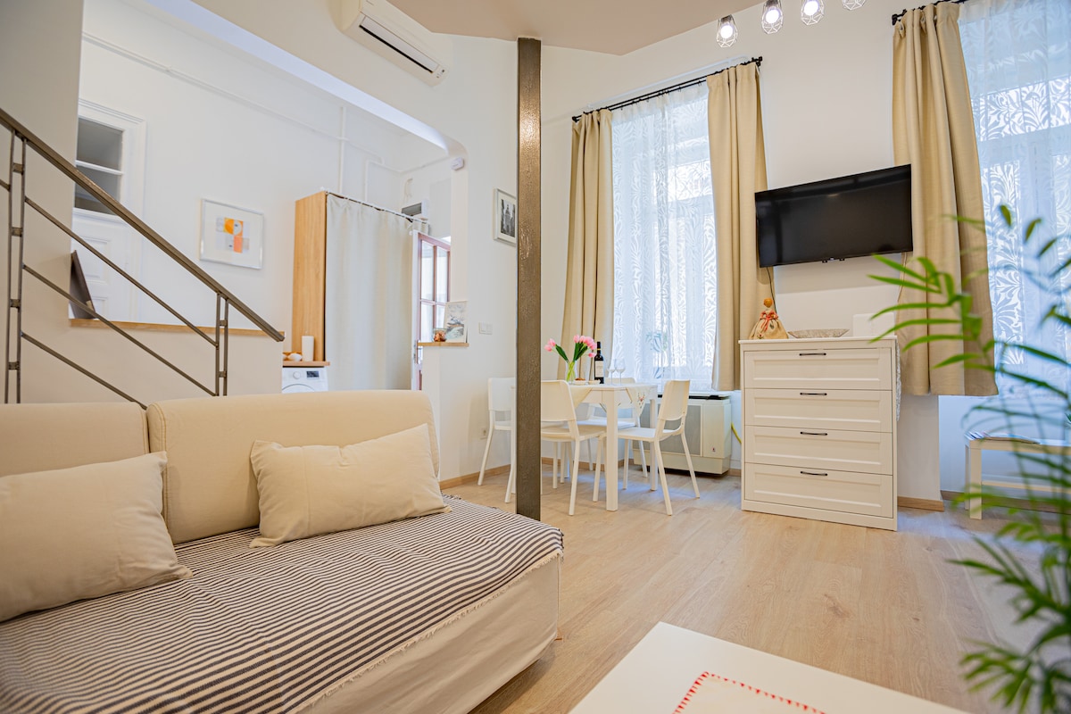 A spacious living area is highlighted by a comfortable sofa and a white dining table, accompanied by four chairs. Natural light filters through sheer curtains covering large windows, enhancing the bright atmosphere. A mounted television is positioned above a stylish dresser.