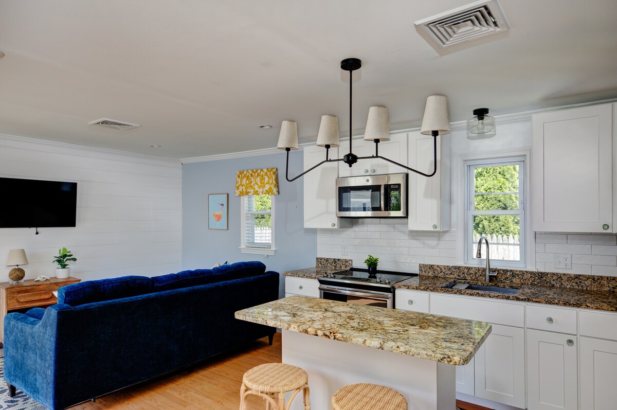 A bright kitchen area features a polished granite countertop and modern appliances. Light-colored cabinetry complements the space, which includes a dining area with two wicker stools. A cozy seating arrangement, highlighted by a navy blue sofa, offers a comfortable spot for relaxation.