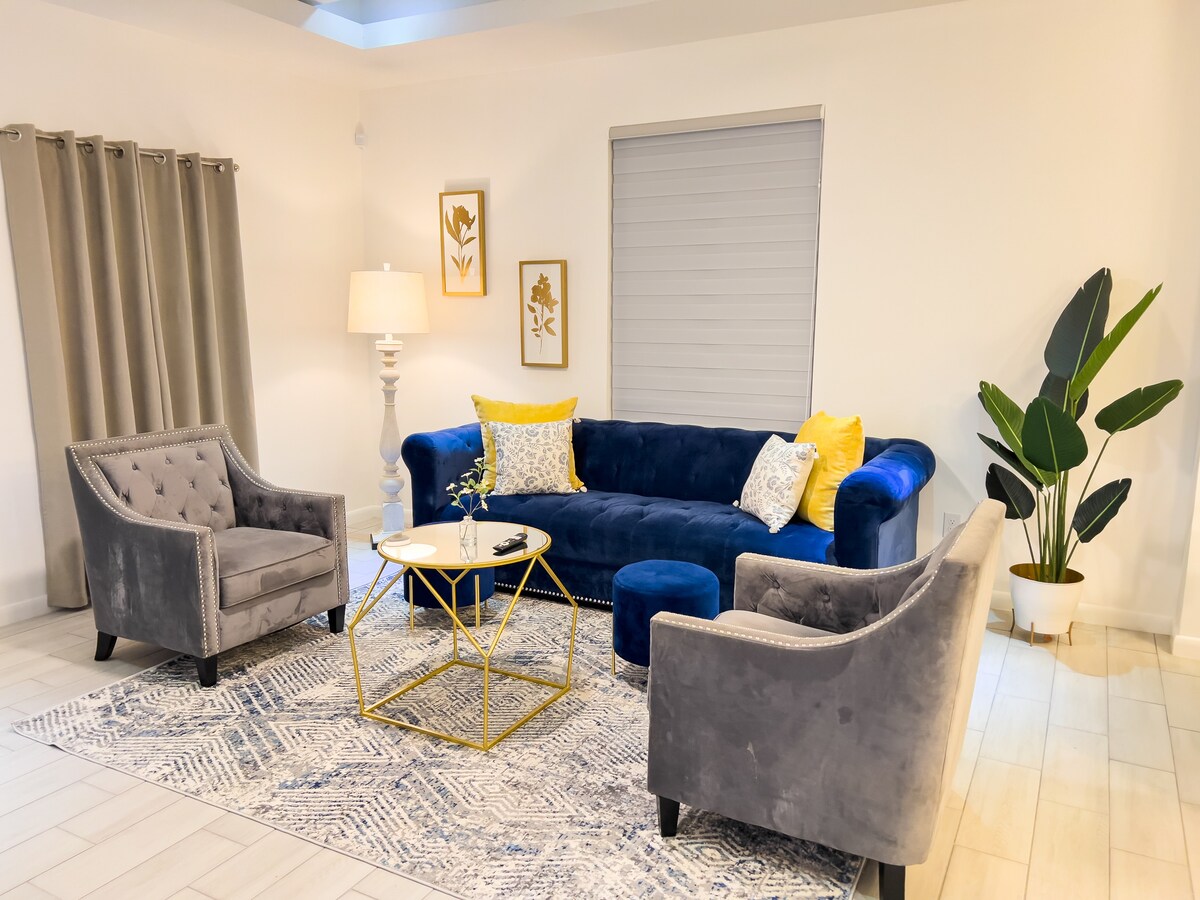A comfortable seating area features a plush blue sofa complemented by yellow and white decorative pillows. Two tufted gray chairs are positioned nearby, creating an inviting atmosphere. A round coffee table with a gold frame sits atop a patterned area rug, while a large potted plant adds a touch of greenery.