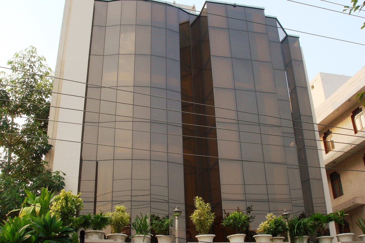 The exterior of the property features a modern facade with large glass windows that reflect the surroundings. Lush greenery is present in planters positioned along the base, contributing to an inviting atmosphere. The building stands prominently against a clear sky.