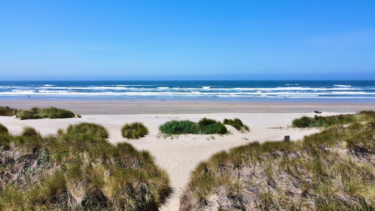 A wide view captures a sandy beach meeting the ocean under a clear blue sky. Gentle waves create a rhythmic pattern along the shore, while grassy dunes frame the pathway to the water, contributing to a serene coastal landscape.