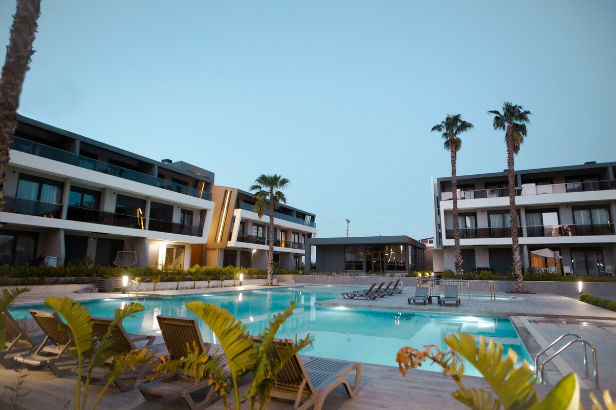 A modern pool area is surrounded by landscaped greenery, featuring lounge chairs arranged around the water. Multiple palm trees frame the site, while contemporary buildings stand in the background under a serene twilight sky.