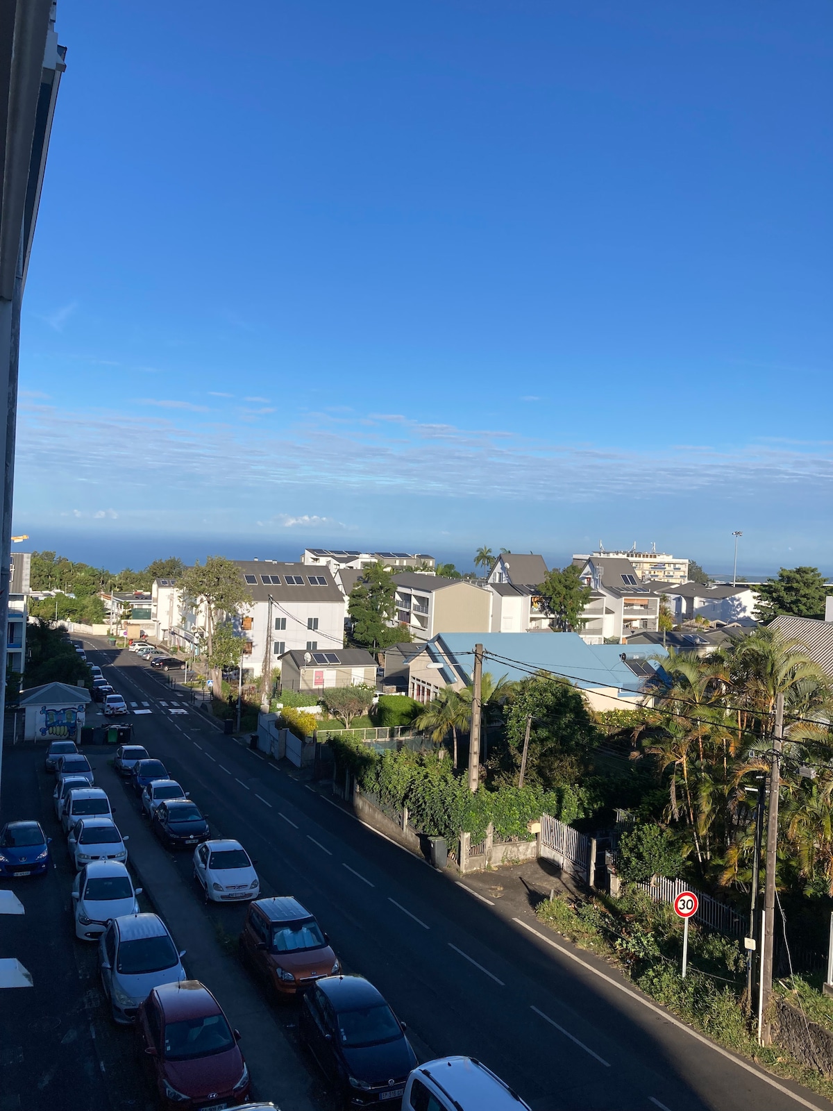 The image captures a panoramic view from the apartment balcony, featuring a clear blue sky and glimpses of the ocean in the distance. Residential buildings and greenery line the street below, with parked cars visible along the road.