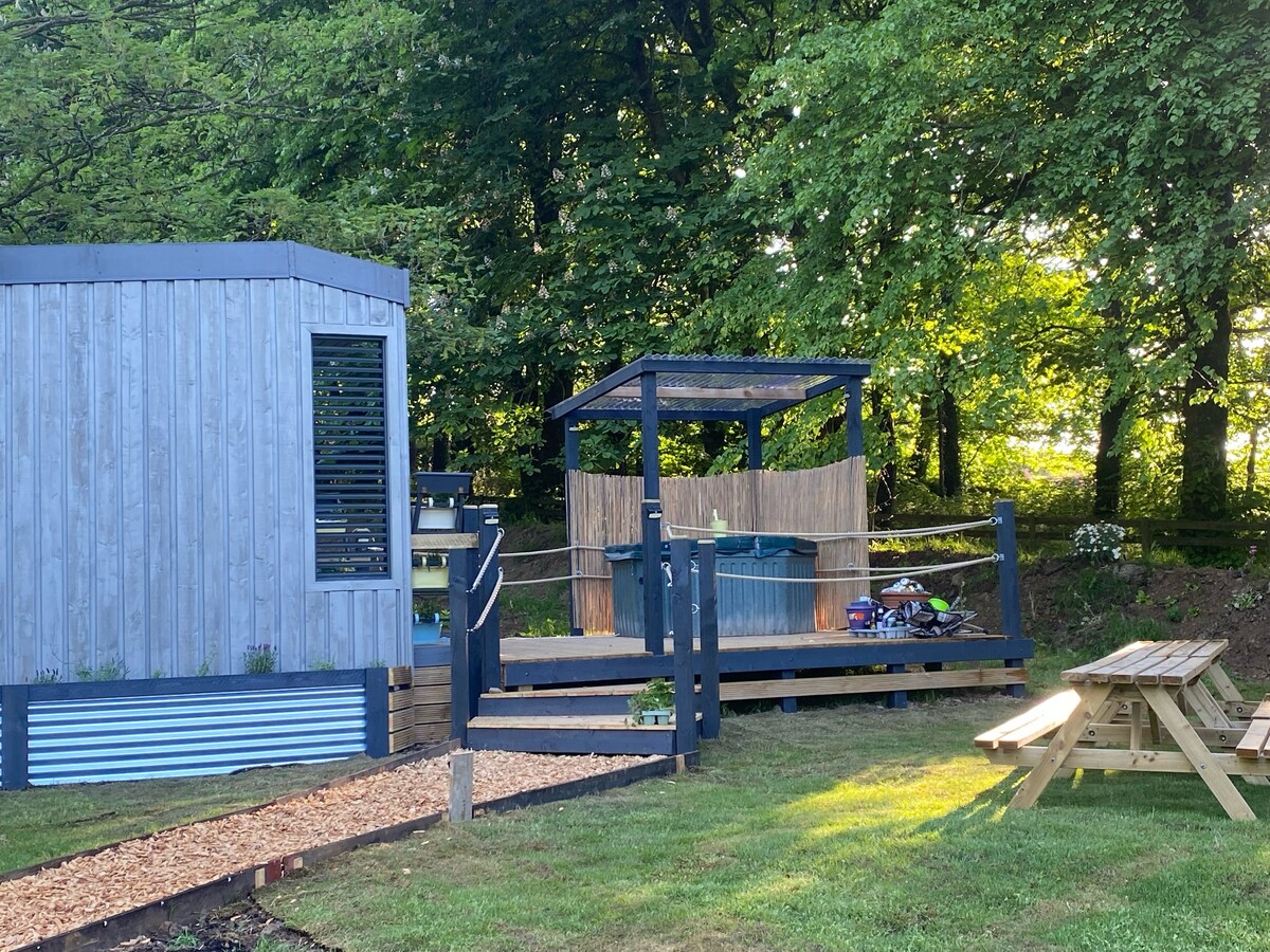 A lodge exterior is shown alongside a wooden deck featuring a hot tub shaded by a gazebo. Lush greenery surrounds the area, with a picnic table positioned on a grassy lawn. Walkways are lined with decorative gravel, leading to the lodge's entrance.