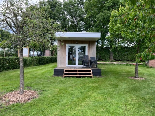 A small cabin is positioned on a grassy area, featuring a wooden deck with steps leading to a glass door. The structure is framed by trees, providing a natural setting that enhances the outdoor space. A table and chairs are situated on the deck.