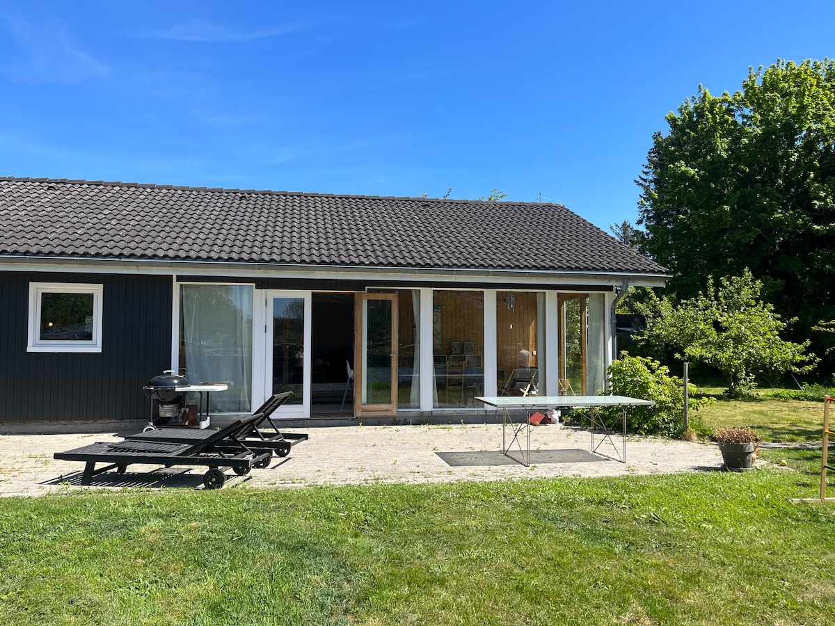 A single-story building features large glass doors that open to a grassy area. Sun loungers and a table are positioned on the patio, with trees providing a natural backdrop. The clear blue sky enhances the spacious outdoor setting.