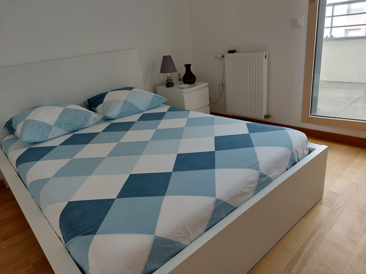 A light-filled bedroom features a spacious bed with a patterned blue and white duvet. A modern bedside table is placed beside the bed, accompanied by a small lamp. Natural light is allowed in through a window, creating a bright atmosphere.