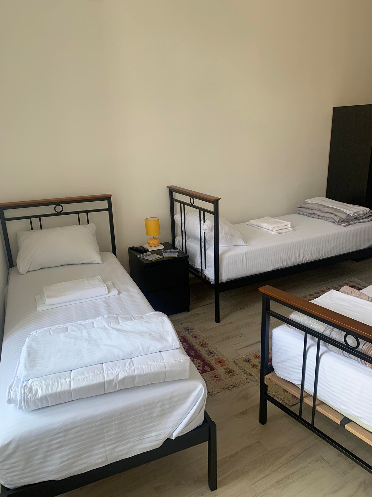 Two twin beds are arranged in a well-lit room, each with fresh linens and a small nightstand beside them. A dark storage cabinet is positioned against one wall, while a rug adds a touch of warmth to the space.