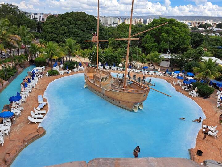 A broad view of an outdoor aquatic area features a large wooden pirate ship in a blue pool. Palm trees and lounge chairs surround the water, providing ample space for relaxation. Bright skies enhance the vibrant atmosphere, inviting guests to enjoy the setting.