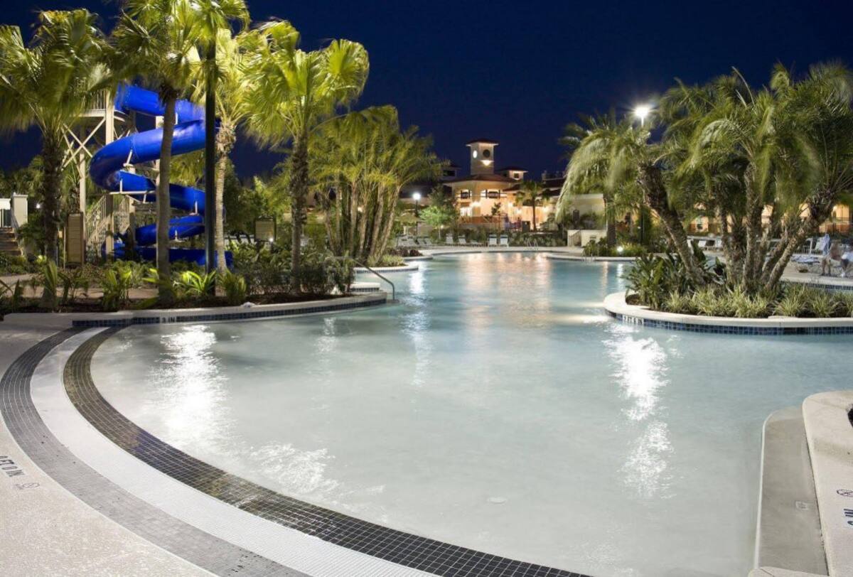 A spacious resort pool is surrounded by palm trees, creating a tropical ambiance. A water slide in bright blue is visible in the background, while illuminated pathways lead around the expansive swimming area, enhancing the inviting atmosphere for relaxation and leisurely activities.