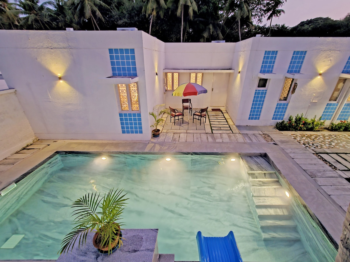 A private pool is surrounded by the elegantly designed villa featuring large windows. Outdoor seating is visible next to the pool, with an umbrella providing shade. Soft lighting enhances the tranquil atmosphere as palm trees are seen in the background.