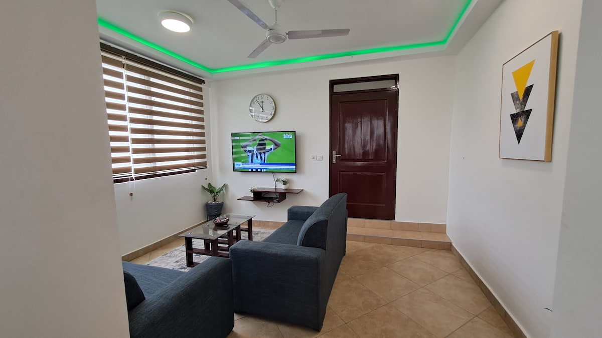 A cozy living area features a dark blue sectional sofa positioned facing a flat-screen television. Natural light filters through stylish blinds, complemented by gentle, ambient lighting on the ceiling. A wall clock and a piece of framed art add a modern touch to the space.