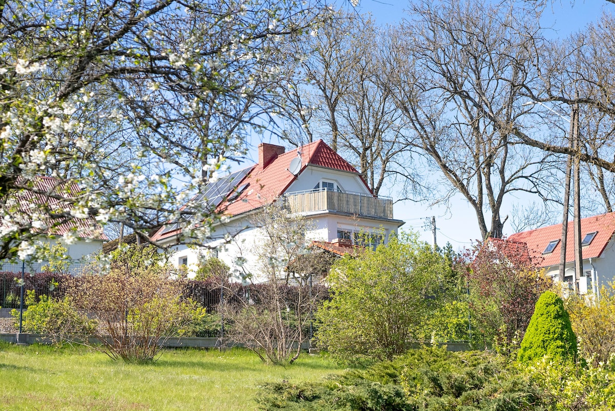 A spacious house with a red-tiled roof is surrounded by lush greenery and flowering trees. The structure features a balcony accessible from the upper level, while the garden exhibits diverse flora, contributing to a serene outdoor setting.