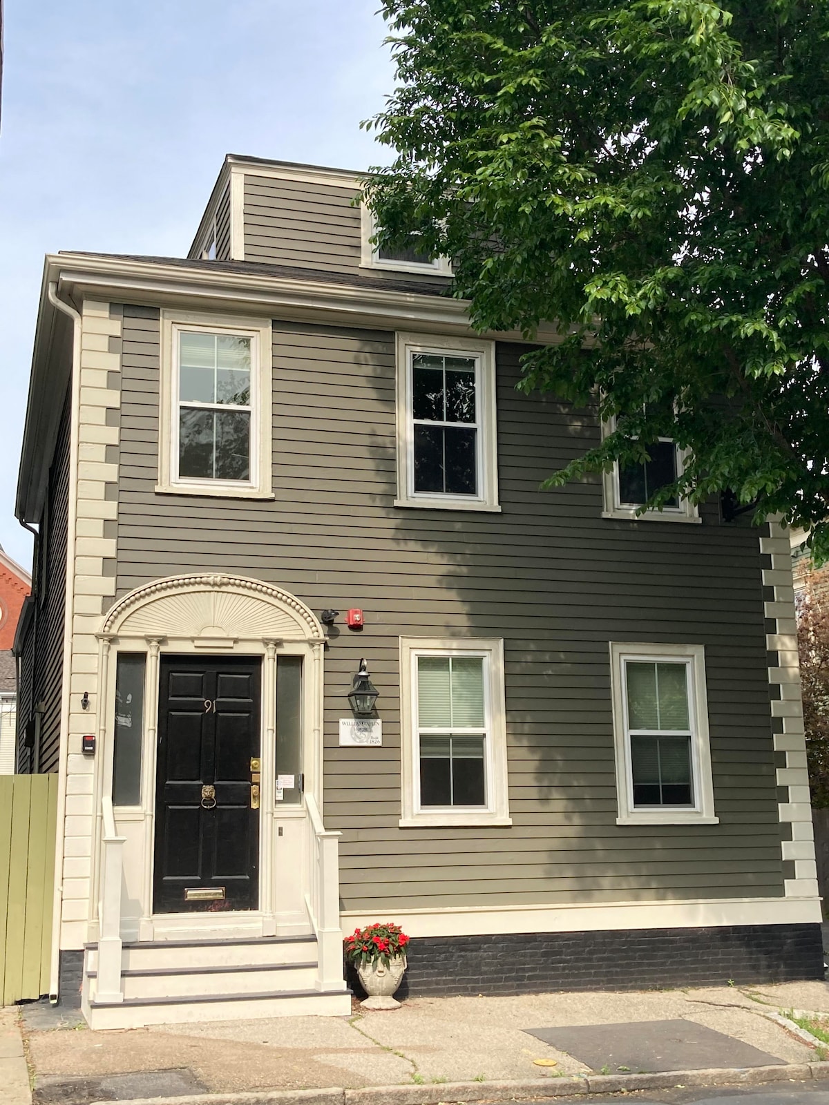 A historic three-story tan and gray building is featured, showcasing symmetrical windows and a decorative entrance. The front door is framed by a classic archway, with a potted plant positioned nearby. Green foliage is visible on a tree in front, enhancing the charm of the exterior.