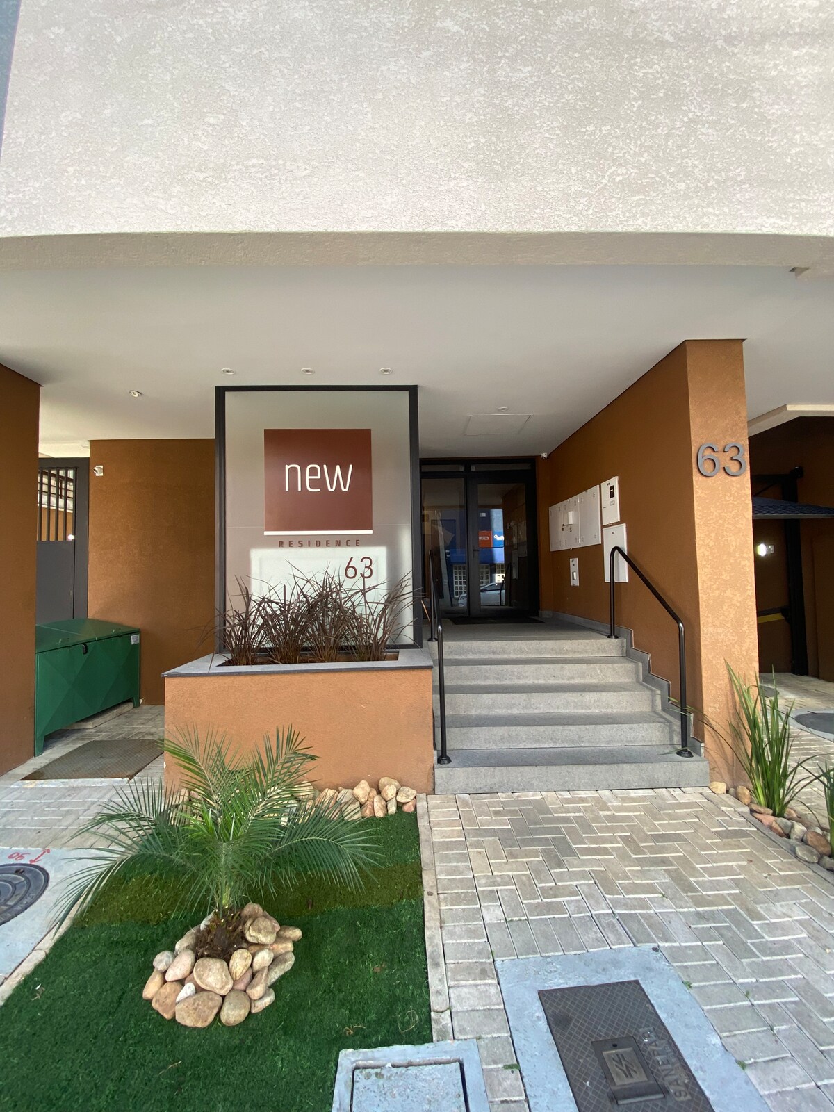 The entrance features a modern design with a large sign displaying 'new' on a brown background. Steps lead up to the entrance, flanked by decorative plants and stones. Smooth pavement and a small patch of grass enhance the welcoming appearance of the property.
