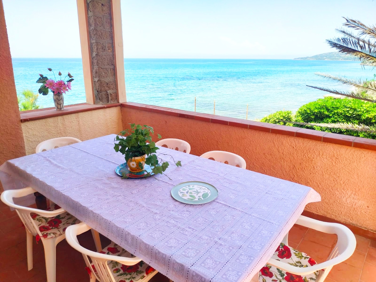A spacious outdoor terrace features a large dining table covered with a light purple tablecloth. White chairs surround the table, and a potted plant sits at the center. In the background, a panoramic view of the sea and shoreline is visible, complemented by greenery.
