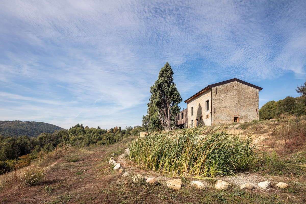 A rustic stone building is set against a backdrop of rolling hills and greenery. The structure features a simple façade with windows, while tall grass and a circular stone arrangement lead the eye to the entrance. The sky is clear with gentle clouds.