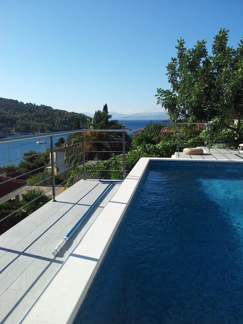 A private pool is set on a terrace, surrounded by lush greenery and offering views of the sea and hills. The clear blue water reflects the sun, creating a serene atmosphere. Simple poolside loungers are positioned for relaxation.