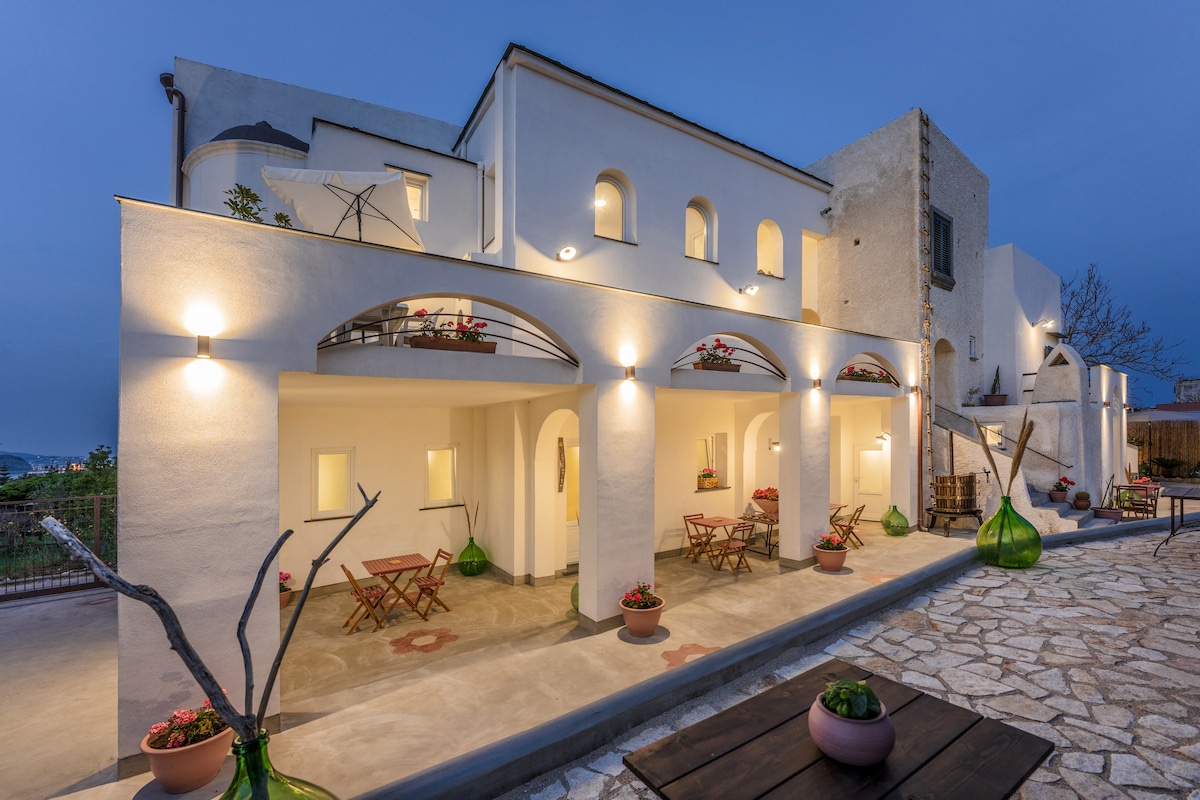 A serene exterior is presented with white-washed walls and arched entryways. Soft lighting highlights the covered terrace, adorned with small tables and chairs. Potted plants are placed throughout, adding a touch of greenery. The smooth stone pathway leads through a tranquil outdoor space.