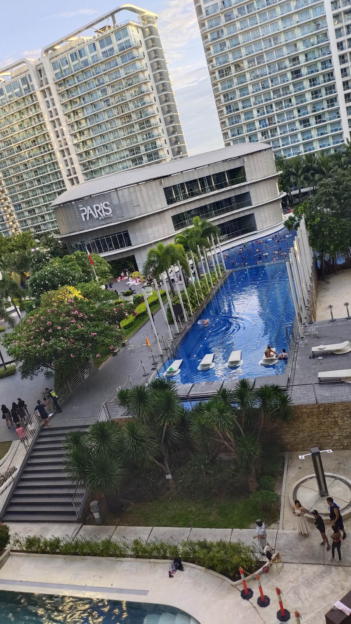An aerial view captures the infinity wave pool at Paris Beach Club, bordered by lush palm trees and modern buildings. Groups of guests can be seen enjoying the water. The pool area features sun loungers and steps leading down to the surrounding greenery.