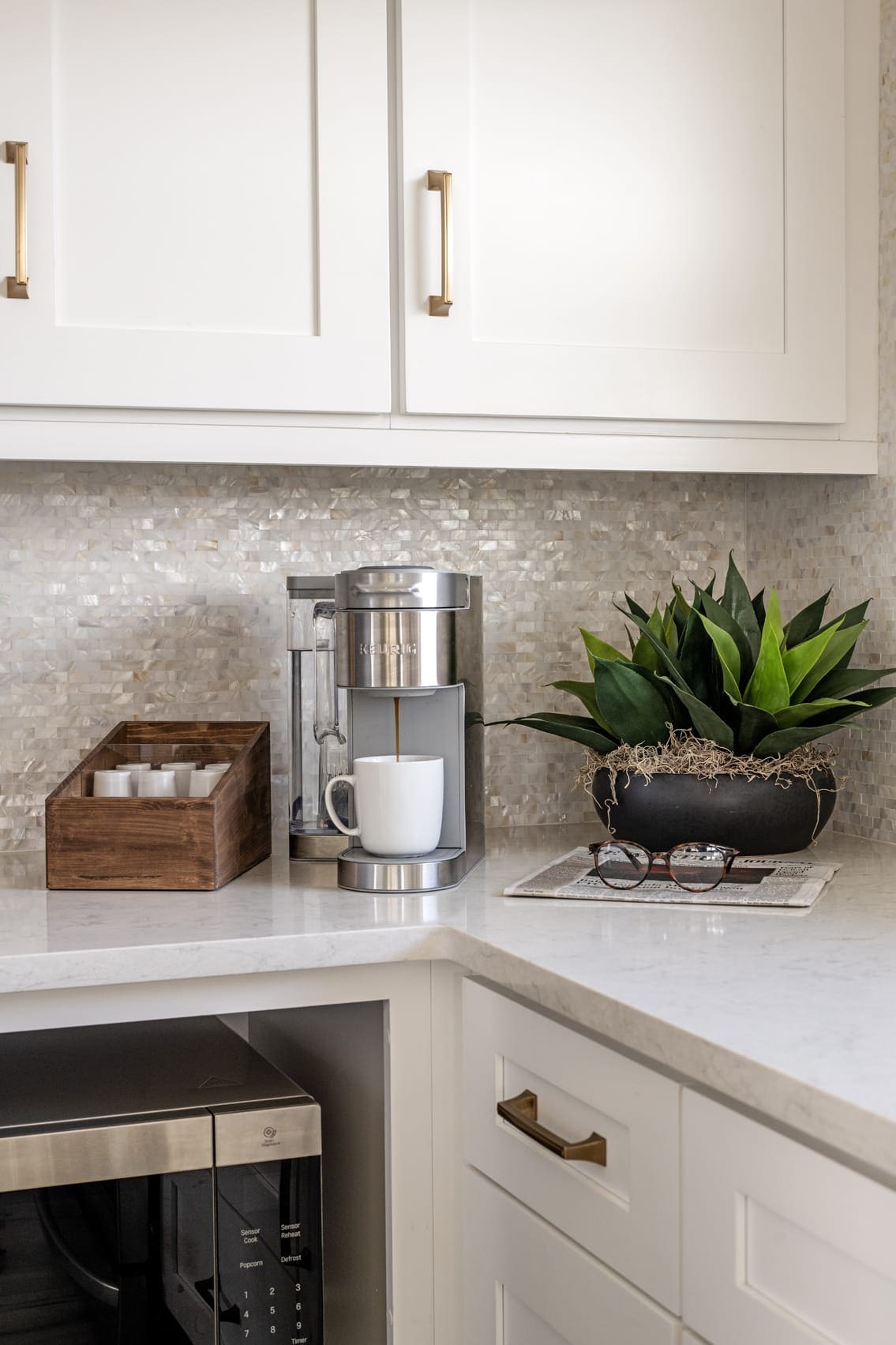 A modern kitchen space features a sleek coffee machine beside a small wooden tray holding sugar and creamers. A potted plant adds a touch of greenery, while light-colored countertops are complemented by a textured backsplash. Lower cabinetry includes a built-in microwave, contributing to a clean and organized aesthetic.