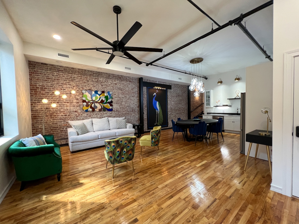 An open living area features a combination of hardwood flooring and exposed brick walls. A large sofa sits alongside colorful accent chairs, while a dining table is positioned nearby. Unique light fixtures and wall art add character to the inviting space.