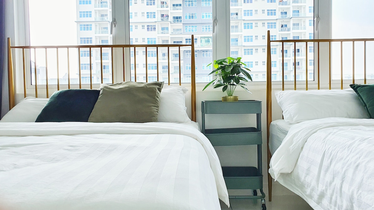 The sleeping area features two queen beds with an arrangement of neutral and deep green pillows. A small, wheeled side table holds a potted plant, while large windows allow natural light to illuminate the space, showcasing a view of the surrounding buildings.